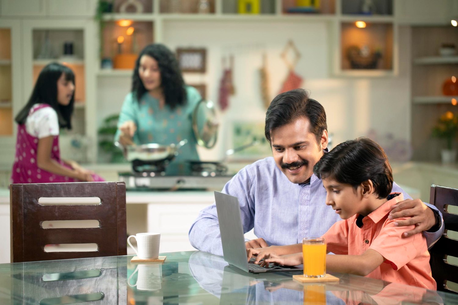An Indian family at home, a father and son share a moment at a laptop while the mother and daughter cook together in the kitchen